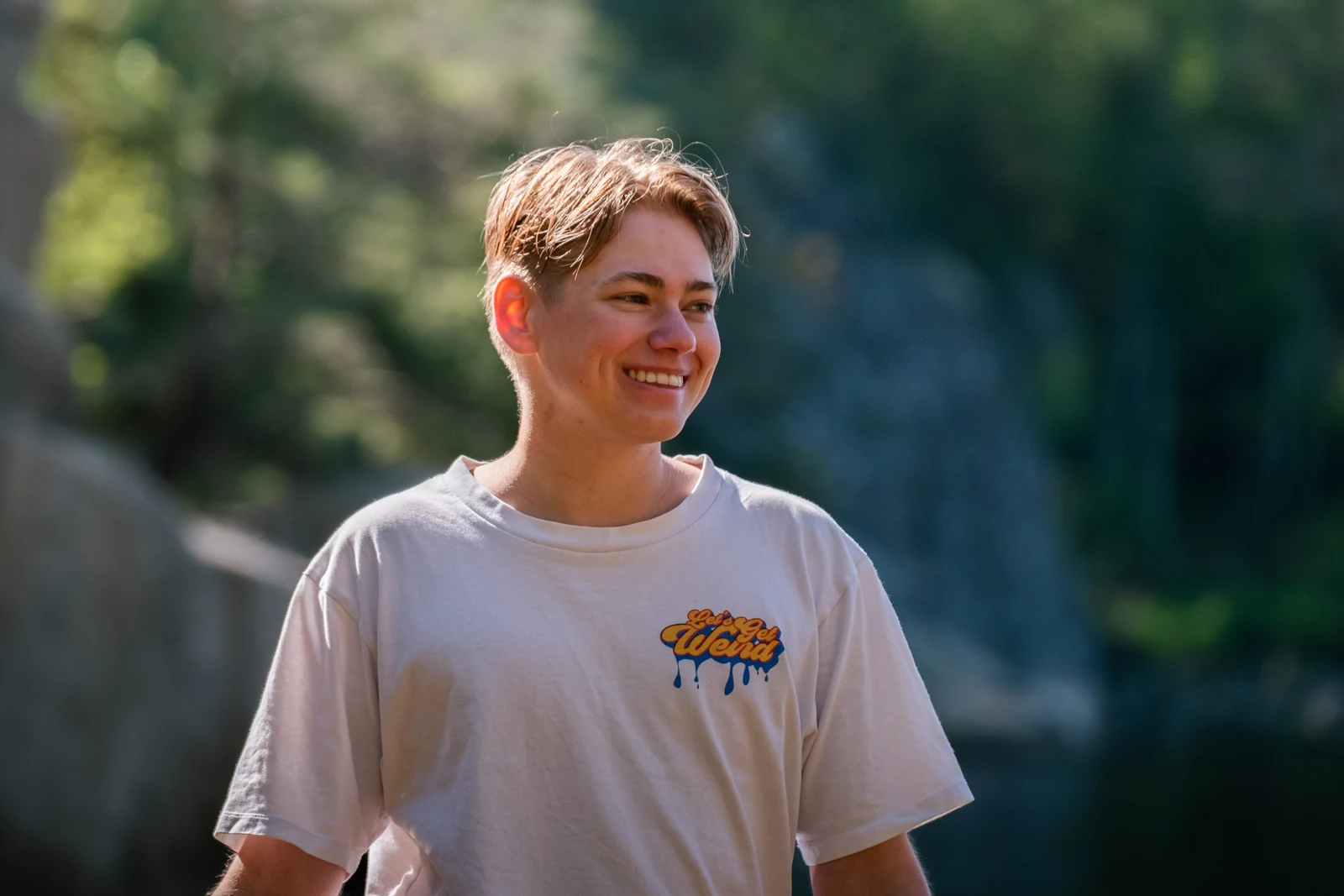 A person in a pale T-shirt smiling outdoors in sunlight