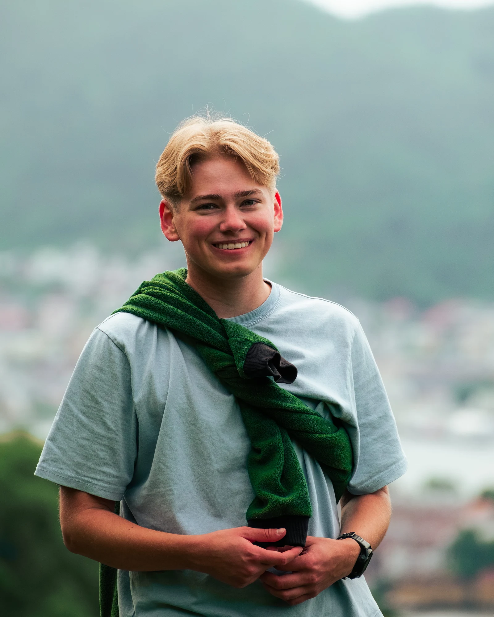 A smiling person with a green sweatshirt over their shoulders and hills behind them