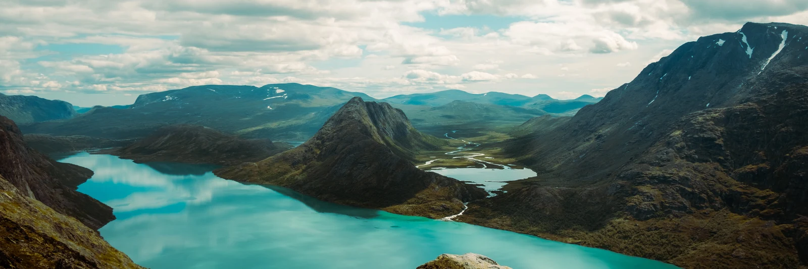 A turquoise lake winding between dark mountain ridges beneath a cloudy sky