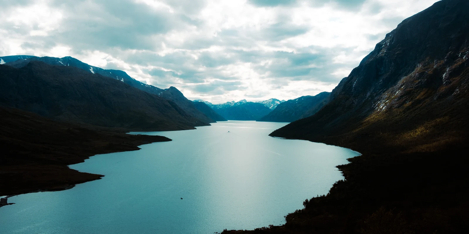 A broad lake winding between steep mountains beneath a bright cloudy sky