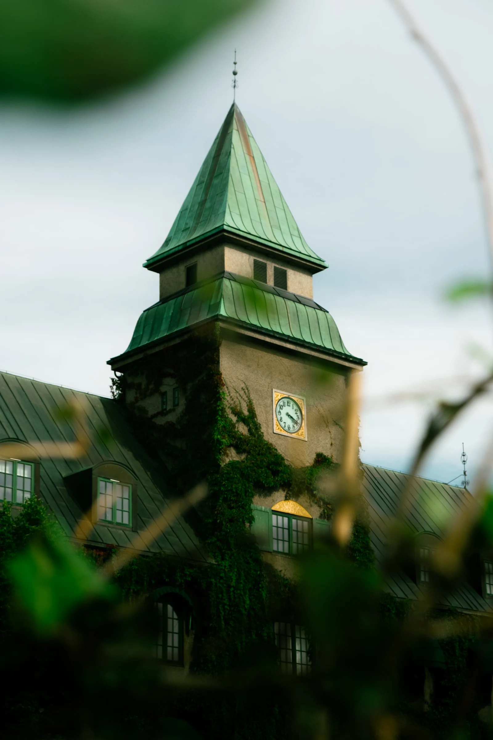 A green-roofed brick clock tower seen through leaves