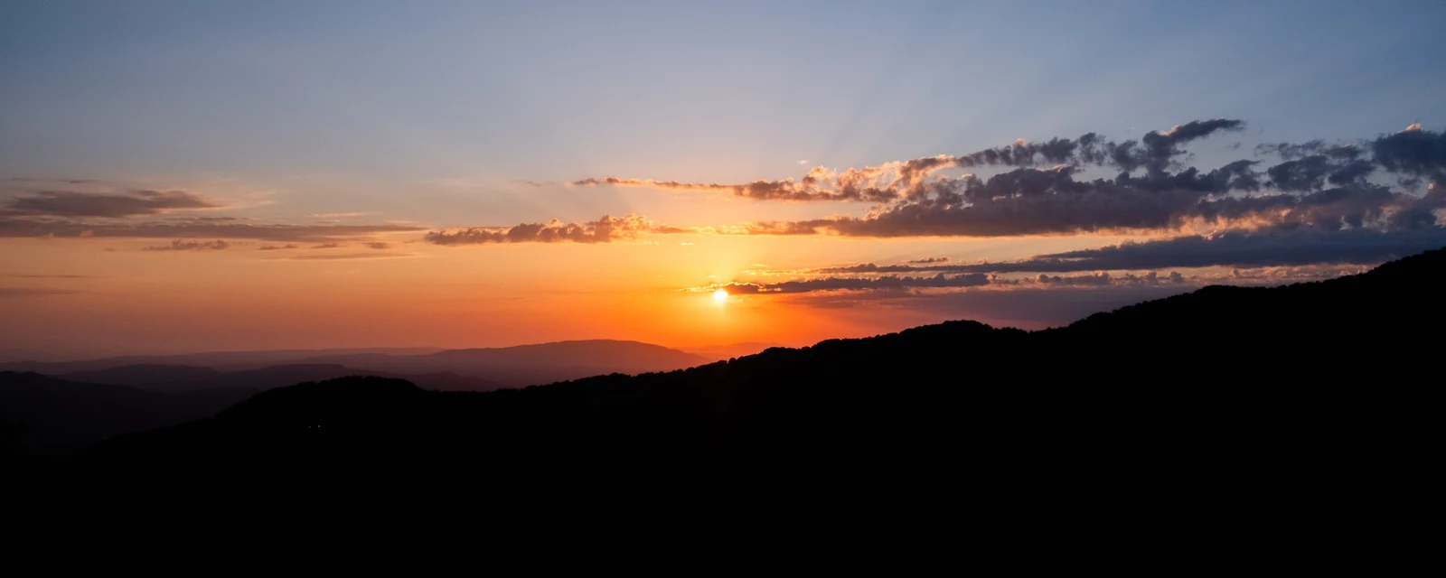 The sun setting beneath blue-grey clouds over a dark hillside