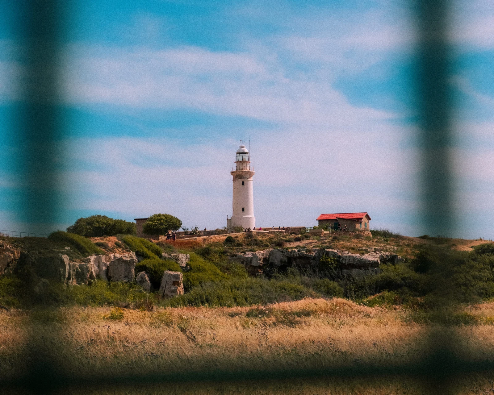 A white lighthouse and small red-roofed building framed by a blurred fence
