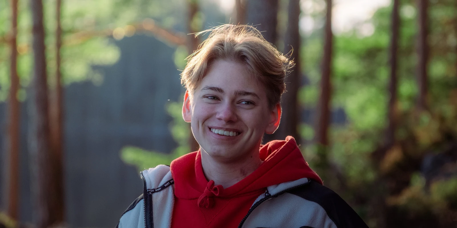 A smiling person in a red hoodie and jacket standing among trees