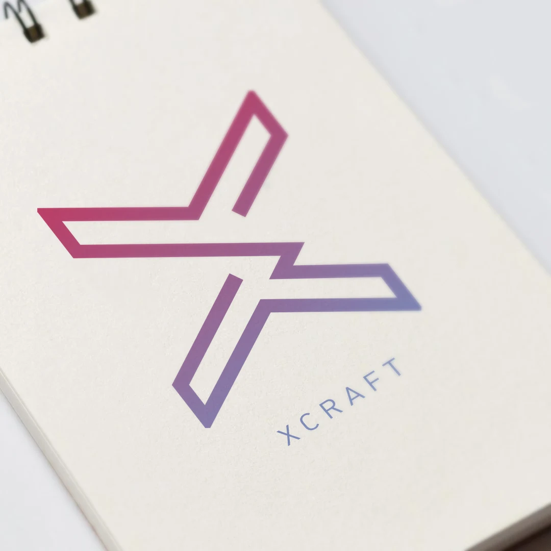 The Xcraft logo in pink and purple printed on a white spiral-bound notebook